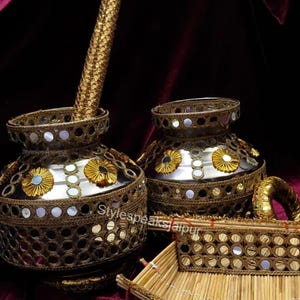 Puede incluir: Dos jarrones ornamentados de metal dorado con intrincados adornos de espejos y flores. Un bol&iacute;grafo dorado a juego est&aacute; en un jarr&oacute;n. Tambi&eacute;n se ve una peque&ntilde;a bandeja rectangular con detalles de espejo y una pulsera dorada. Los art&iacute;culos est&aacute;n sobre un fondo burdeos oscuro.