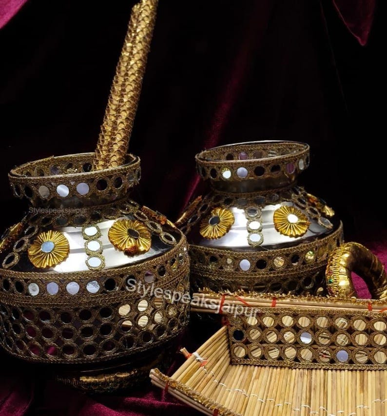Puede incluir: Dos jarrones ornamentados de metal dorado con intrincados adornos de espejos y flores. Un bol&iacute;grafo dorado a juego est&aacute; en un jarr&oacute;n. Tambi&eacute;n se ve una peque&ntilde;a bandeja rectangular con detalles de espejo y una pulsera dorada. Los art&iacute;culos est&aacute;n sobre un fondo burdeos oscuro.