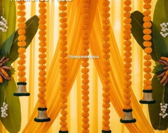 May include: A decorative arrangement featuring strings of orange marigold flowers, hanging in front of a yellow curtain. The garlands have green and gold embellishments. The image includes the text "Reference Photo" and "Stylespeaksjaipur".