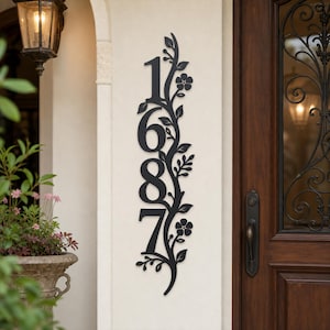 May include: A vertical black metal house number sign displaying the digits 1687, integrated with a decorative vine pattern featuring leaves and small flowers. The sign is mounted on a light-colored wall next to a dark wooden door with glass.