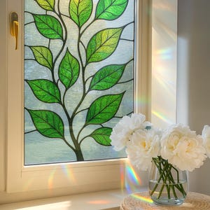 Puede incluir: Una ventana con un diseño de vidriera que presenta una rama de árbol con hojas verdes. Un jarrón de flores blancas se encuentra sobre un taburete tejido cerca de la ventana, con la luz solar creando un efecto arcoíris.