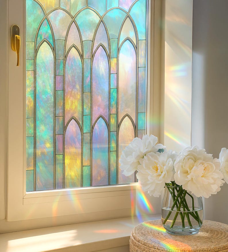 May include: A window with a stained glass design featuring a spectrum of colors, including blue, yellow, and pink. A bouquet of white peonies in a clear glass vase sits on a woven stool near the window.