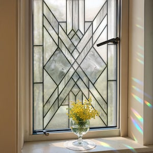 Könnte beinhalten: Ein Fenster mit einem geometrischen Buntglasdesign, das klare und mattierte Glasscheiben in schwarzen Linien umrahmt. Ein kleines Glas mit gelben Blumen steht auf der Fensterbank, darunter eine weiße Spitze.