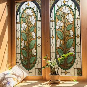Op de afbeelding: Twee glas-in-loodramen met een bloemenontwerp met witte bloemen en groene bladeren. De ramen zijn omlijst met hout en geplaatst in een houten muur. Een kleine potplant staat op de vensterbank, samen met een beige kussen.
