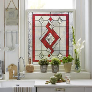 May include: A window with a stained glass panel featuring a geometric design in red and clear glass. The window is framed in red. Small potted plants sit on the windowsill, along with a sink and chrome faucet.