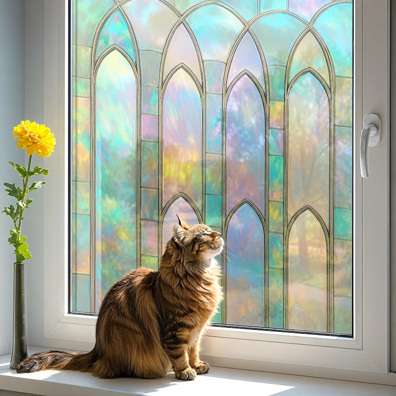 May include: A brown tabby cat sits on a white windowsill, gazing upwards. A stained-glass window with arched panels provides a colorful backdrop. A yellow flower in a slender vase is to the left of the window.