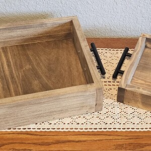 Puede incluir: Dos bandejas de madera para servir con acabado natural y asas negras. Las bandejas rectangulares tienen laterales elevados y est&aacute;n colocadas sobre un camino de mesa de encaje decorativo. La bandeja m&aacute;s grande mide unos 40 cm de largo, y la m&aacute;s peque&ntilde;a, unos 25 cm.