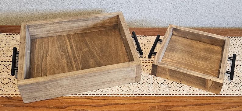 Puede incluir: Dos bandejas de madera para servir con acabado natural y asas negras. Las bandejas rectangulares tienen laterales elevados y est&aacute;n colocadas sobre un camino de mesa de encaje decorativo. La bandeja m&aacute;s grande mide unos 40 cm de largo, y la m&aacute;s peque&ntilde;a, unos 25 cm.