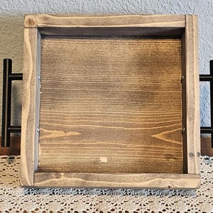 Puede incluir: Una bandeja de madera cuadrada con acabado te&ntilde;ido. La bandeja tiene un borde elevado y asas de metal negro a cada lado. La veta de la madera es visible y la bandeja parece hecha a mano.