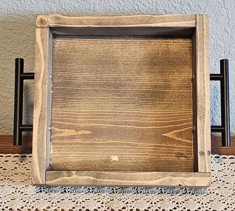 Puede incluir: Una bandeja de madera cuadrada con acabado te&ntilde;ido. La bandeja tiene un borde elevado y asas de metal negro a cada lado. La veta de la madera es visible y la bandeja parece hecha a mano.