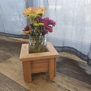 Könnte beinhalten: Ein hölzerner Pflanzkasten mit einem Glasgefäß voller bunter Blumen. Die Blumen sind gelb, grün und lila. Der Pflanzkasten ist aus hellem Holz gefertigt und steht auf einem Holzboden.