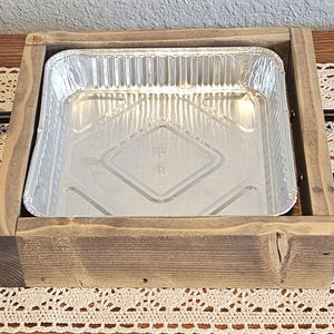 Puede incluir: Una bandeja de madera con una bandeja rectangular de papel de aluminio. La bandeja es de madera marr&oacute;n claro y tiene asas de metal negro a cada lado. La bandeja de aluminio est&aacute; vac&iacute;a y tiene un patr&oacute;n de diamantes en la parte inferior.