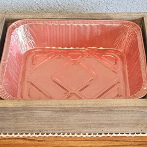 Puede incluir: Una bandeja de madera con asas negras contiene una bandeja rectangular desechable para hornear de papel de aluminio rosa. La bandeja es de madera de color claro y se encuentra sobre un mantel de encaje blanco.