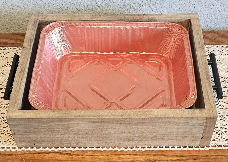 Puede incluir: Una bandeja de madera con asas negras contiene una bandeja rectangular desechable para hornear de papel de aluminio rosa. La bandeja es de madera de color claro y se encuentra sobre un mantel de encaje blanco.