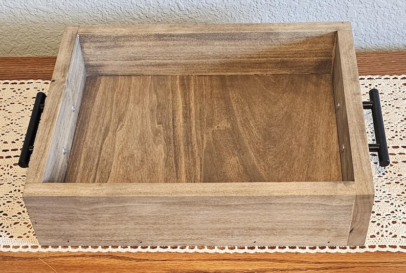 Puede incluir: Una bandeja rectangular de madera con un acabado de vetas naturales. Cuenta con asas de metal negro en cada lado. La bandeja est&aacute; dise&ntilde;ada con una est&eacute;tica sencilla y limpia, adecuada para servir o exhibir.