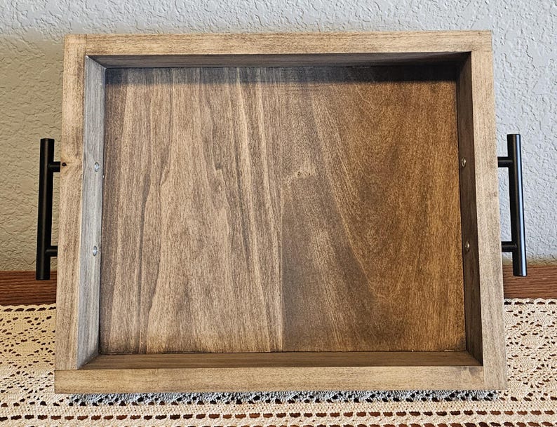 Puede incluir: Una bandeja de madera rectangular con acabado te&ntilde;ido. La bandeja tiene un borde elevado y asas de metal negro en cada extremo. La veta de la madera es visible.