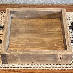 Puede incluir: Una bandeja cuadrada de madera con acabado te&ntilde;ido y asas de metal negro. La bandeja tiene un borde elevado y un patr&oacute;n de vetas de madera natural. Las asas est&aacute;n fijadas a los lados.