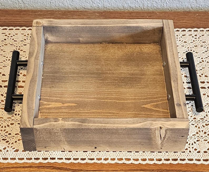 Puede incluir: Una bandeja cuadrada de madera con acabado te&ntilde;ido y asas de metal negro. La bandeja tiene un borde elevado y un patr&oacute;n de vetas de madera natural. Las asas est&aacute;n fijadas a los lados.