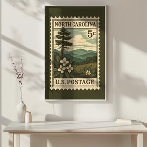 May include: A framed wall art print featuring a vintage-style postage stamp design of North Carolina. The illustration depicts a tall pine tree, white rhododendron flowers, and rolling blue mountains under a cloudy sky with the text North Carolina 5c.