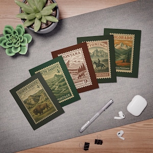 May include: A collection of vintage-style postcards featuring illustrations of U.S. states, including North Dakota, South Dakota, Montana, Wyoming, and Colorado. Each postcard has a postage stamp design with scenic landscapes and text. The postcards are arranged on a gray surface.