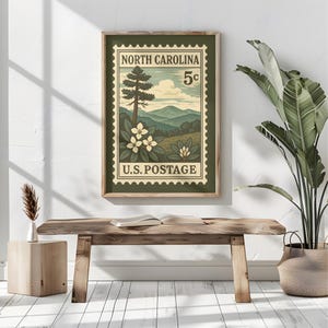 May include: A framed rectangular wall art print featuring a vintage-style postage stamp design. The graphic depicts a North Carolina landscape with mountains, a tall pine tree, and white flowers. Text reads NORTH CAROLINA, 5c, and U.S. POSTAGE.
