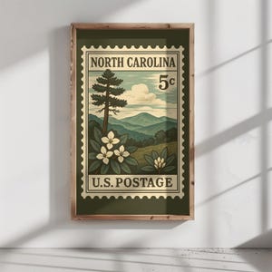 May include: A framed wall art print featuring a vintage-style postage stamp design of North Carolina. The illustration depicts a tall pine tree, white rhododendron flowers, and rolling blue mountains under a cloudy sky with the text 5c U.S. POSTAGE.
