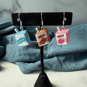 May include: Three novelty earrings shaped like milk cartons in blue, orange, and pink. Each carton has a different design and the word "MILK" printed on it. The earrings are displayed on a black earring stand.