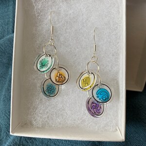 May include: A pair of silver earrings with clear plastic charms featuring different colored symbols. The symbols are green, yellow, blue, purple, and white.