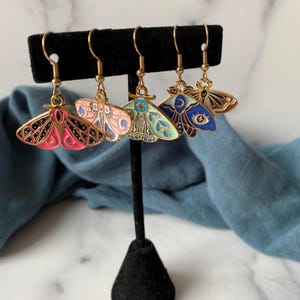 May include: Five pairs of gold-tone earrings with colorful enamel moth designs. The moths have different patterns and colors, including pink, blue, green, and black.