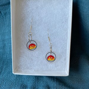 May include: A pair of dangle earrings featuring a cartoon design of a face with yellow and red colors. The earrings are displayed inside a white box with a blue background.