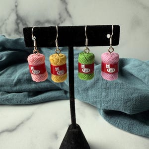 May include: Four pairs of yarn ball earrings in pink, yellow, and green, each with a red band and a cartoon face. The earrings are displayed on a black jewelry stand against a blue fabric backdrop.