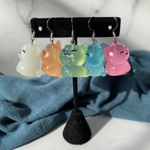 May include: Five pairs of cat-shaped earrings in various colors: white, orange, green, blue, and pink. Each earring features a smiling cat design with silver-colored ear wires. The earrings are displayed on a black earring stand.