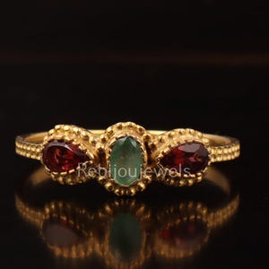 Puede incluir: Un anillo de oro con tres piedras preciosas: una piedra verde ovalada en el centro, flanqueada por dos piedras rojas en forma de pera. El anillo tiene una banda texturizada y engastes decorativos.