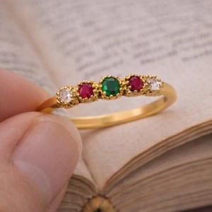 Puede incluir: Un anillo de oro con una fila de piedras preciosas. El anillo tiene una piedra verde central flanqueada por piedras rojas y blancas. El anillo se sostiene sobre un libro abierto, mostrando el diseño y los colores del anillo.