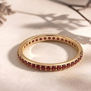 Puede incluir: Un anillo dorado con una banda de pequeñas gemas rojas redondas. El anillo se encuentra sobre una superficie clara, con sombras suaves en el fondo. El anillo es una banda de eternidad completa, con gemas que rodean todo el anillo.