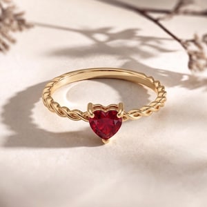 Könnte beinhalten: Ein goldener Ring mit einem geflochtenen Band und einem herzförmigen, roten Edelstein. Der Edelstein ist der Mittelpunkt und in einer zarten Vier-Zacken-Fassung gefasst. Der Ring wird auf einer neutralen Oberfläche mit weichen Schatten präsentiert.