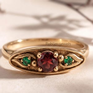Può includere: Anello dorato con una pietra rossa centrale e due pietre verdi. L'incastonatura è ornata da un motivo floreale. Le pietre sono rotonde. Un gioiello.