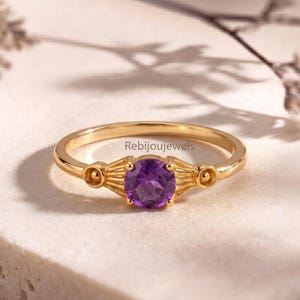 May include: A gold-colored ring with a round, deep purple gemstone at the center. The ring has a delicate band and decorative elements on either side of the gemstone. The text "Rebijoujewels" is visible on the ring.