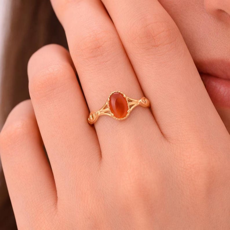 May include: A gold ring featuring an oval, orange-red gemstone. The ring has a delicate, openwork design with decorative elements on the sides. The gemstone is set in a detailed bezel.