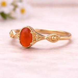May include: A gold ring featuring an oval, orange-red gemstone. The ring has an ornate design with decorative elements on the band. The gemstone is set in a detailed bezel setting. The ring is likely made of gold.
