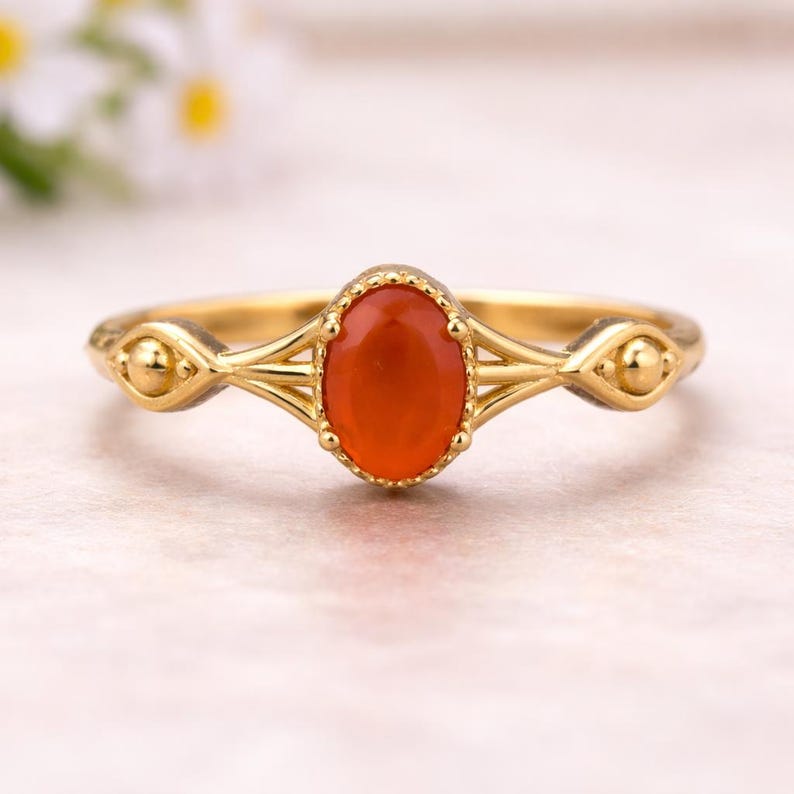 May include: A gold-colored ring featuring an oval, orange-red gemstone. The ring has a delicate design with decorative elements on the band and around the gemstone. The gemstone is approximately 0.5 inches in length. This ring is a piece of jewelry.