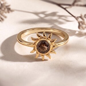May include: A gold ring with a sunburst design. The ring features a round, brown gemstone at the centre of the sun. The sunburst has pointed rays. The ring is set against a neutral background.