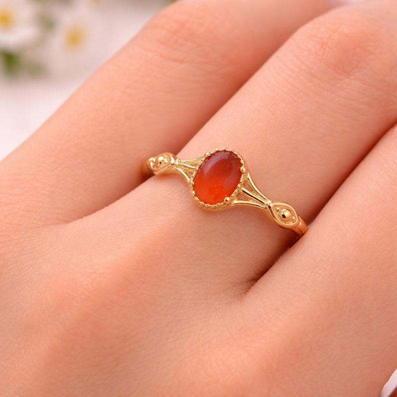 May include: A gold ring featuring an oval, reddish-orange gemstone. The ring has a delicate, ornate design with small decorative elements on the band. The gemstone is set in a gold bezel, and the ring is shown on a person's finger.