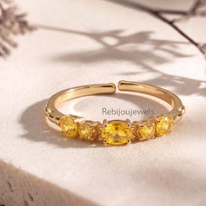 Peut inclure: Une bague dorée ornée de pierres précieuses jaunes. La bague est ouverte et ornée d'une pierre ovale centrale flanquée de pierres rondes plus petites. Le texte "Rebijoujewels" est visible sur la bague.