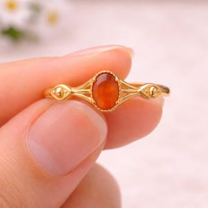 May include: Gold ring featuring an oval, orange gemstone. The ring has a delicate, filigree design with small, round accents on either side of the gemstone. The ring is held by a person.