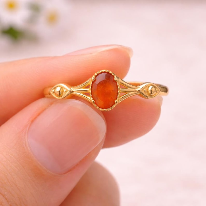 May include: Gold ring featuring an oval, orange gemstone. The ring has a delicate, filigree design with small, round accents on either side of the gemstone. The ring is held by a person.