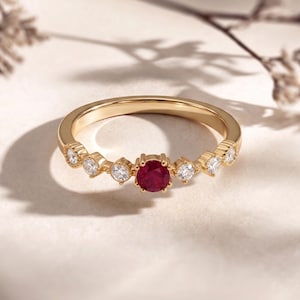 Könnte beinhalten: Ein goldener Ring mit einem zentralen runden Rubin und sechs kleineren, klaren Edelsteinen. Das Band des Rings hat dekorative Details. Die Edelsteine sind in einem goldenen Band gefasst und schaffen ein klassisches und elegantes Design.