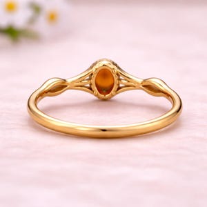 May include: A gold ring with an oval, amber-colored gemstone. The ring has a delicate, openwork design with a slender band and decorative accents on the shoulders. The gemstone is set in a bezel setting.