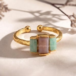 May include: A gold-toned adjustable ring featuring a central rectangular lavender stone flanked by two rectangular light green stones. The ring has a textured band and the brand name "Rebijoujewels" is visible.