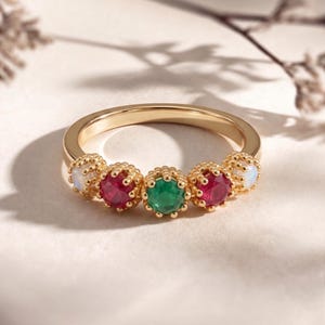 Könnte beinhalten: Ein goldener Ring mit fünf Edelsteinen: zwei roten Rubinen, einem grünen Smaragd und zwei Opalen. Die Edelsteine sind in verzierten, perlenbesetzten Fassungen gefasst. Der Ring wird auf einer hellen Oberfläche gezeigt.